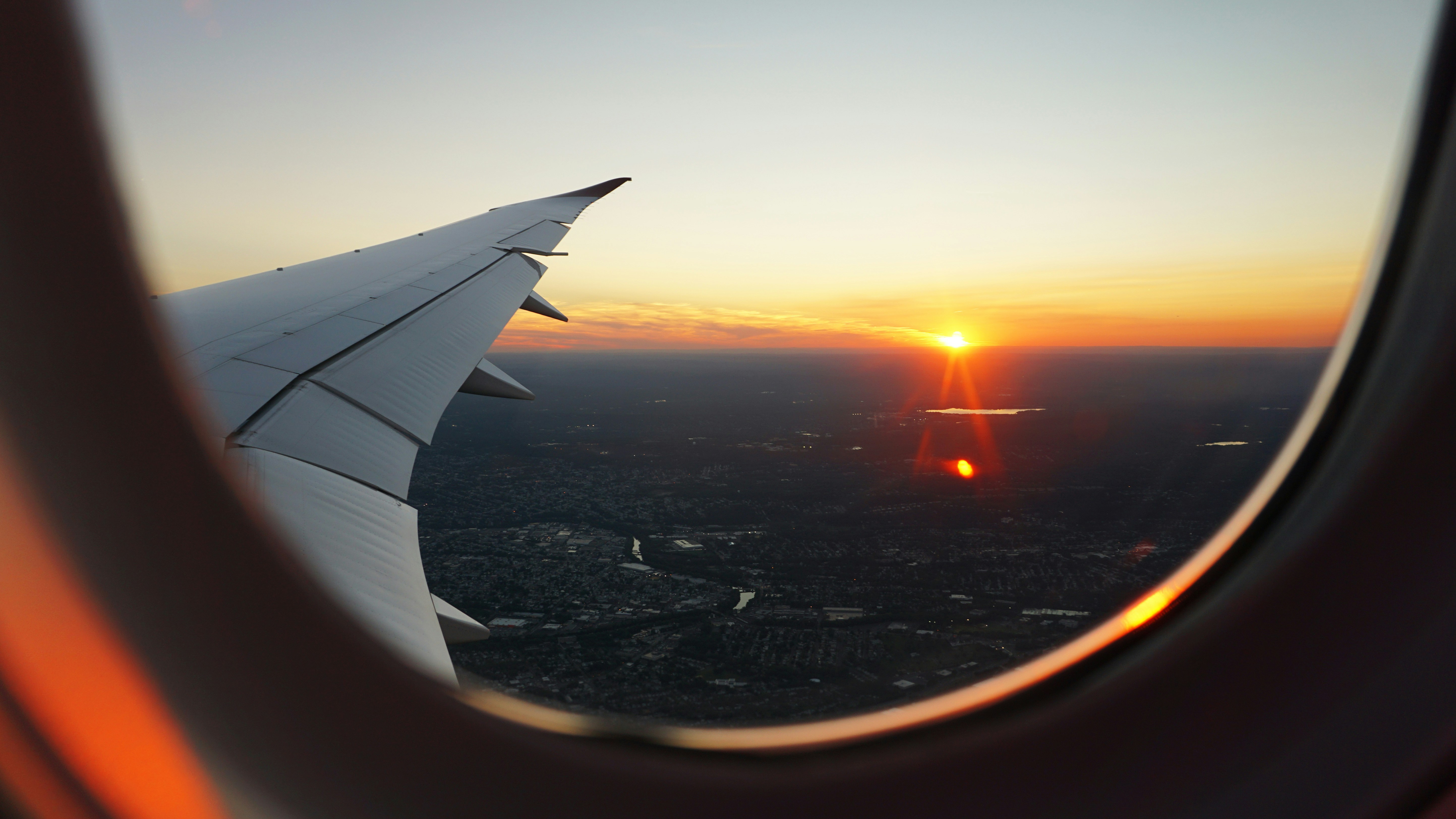 View from an airplane window during sunset, with the airplane wing visible and a landscape below with scattered lights. The sky is painted with warm hues of orange, pink, and purple as the sun sets, reflecting on a distant body of water.